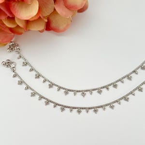 May include: Two silver anklets with delicate floral and teardrop charms. The anklets are displayed on a white surface with a blurred background of orange and pink flower petals. The anklets are connected by a chain.