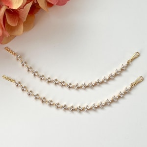 May include: Two delicate gold-toned hair chains with a leaf-like design, adorned with small, clear crystals. Each chain has a hook at one end and a loop at the other. The chains are displayed on a white surface, with a blurred floral element in the background.