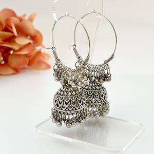 May include: Silver-toned hoop earrings with dangling bell-shaped charms. The earrings have intricate filigree detailing and small, round beads. The hoops are displayed on a clear stand, with a blurred floral arrangement in the background.