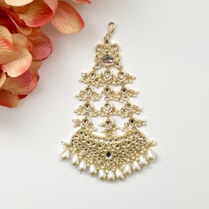 Może przedstawiać: Zdobiona ozdoba do włosów w kolorze złotym i białym z perłowymi akcentami. Ozdoba ma kształt łzy i posiada duży, okrągły, lustrzany element centralny.