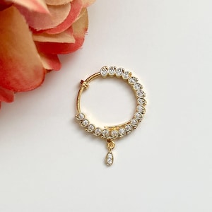 May include: Gold nose ring with a teardrop-shaped pendant and a circle of small, clear stones.