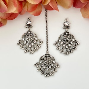May include: Silver-toned jewelry set featuring a pair of earrings and a headpiece. The earrings and headpiece have a heart-shaped design with intricate detailing and dangling accents. The set is displayed against a white background with floral accents.