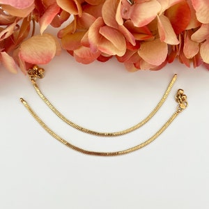 May include: Two gold anklets with a delicate chain design and small round charms. The anklets are laid out on a white surface.