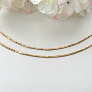Könnte beinhalten: Zwei goldfarbene, flache Schlangenkettchen vor weißem Hintergrund, mit weißen und rosa Blüten im oberen Teil des Bildes. Die Ketten sind dünn und haben ein strukturiertes, schuppenartiges Aussehen, was auf ein filigranes Design hindeutet.