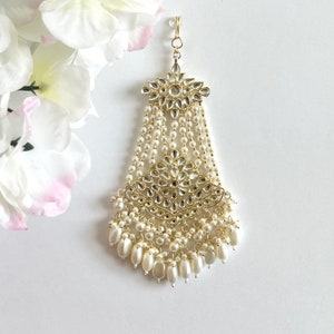 Op de afbeelding: Een gouden haaraccessoire met witte parels en een groot, versierd bloemenmotief. Het accessoire heeft een haak om aan het haar te bevestigen.