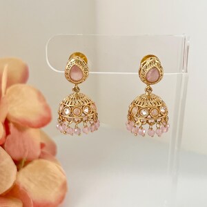 May include: A pair of gold-toned earrings with a teardrop-shaped pink gemstone at the top and a bell-shaped design. The earrings feature intricate detailing and small pink beads hanging from the bottom.