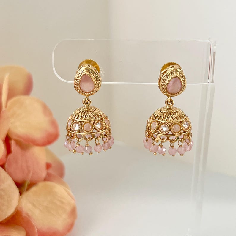 May include: A pair of gold-toned earrings with a teardrop-shaped pink gemstone at the top and a bell-shaped design. The earrings feature intricate detailing and small pink beads hanging from the bottom.