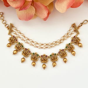 May include: An ornate gold-coloured jewellery piece with detailed pendants, adorned with red and green accents, linked by delicate pearl chains. Small, dangling gold spheres complete the design.