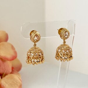 May include: A pair of gold-colored earrings with a teardrop-shaped top and a bell-shaped bottom. The earrings are adorned with small, clear gemstones and dangling accents. The earrings are displayed on a clear stand against a soft, neutral background.