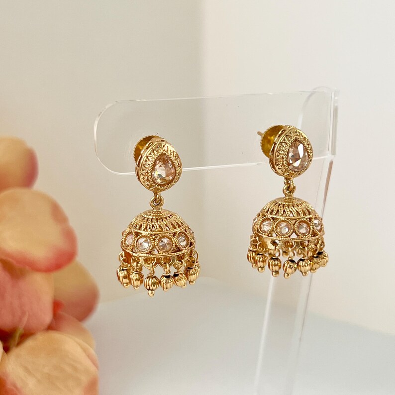 May include: A pair of gold-colored earrings with a teardrop-shaped top and a bell-shaped bottom. The earrings are adorned with small, clear gemstones and dangling accents. The earrings are displayed on a clear stand against a soft, neutral background.