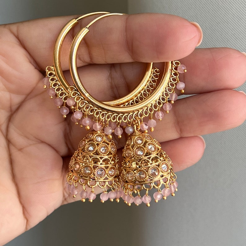 May include: Gold hoop earrings with pink bead accents and dangling jhumka charms. The jhumkas are decorated with gold filigree and clear stones.