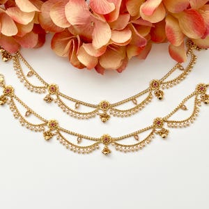 Pode incluir: Duas tornozeleiras douradas com detalhes de flores cor-de-rosa e enfeites de p&eacute;rolas. As tornozeleiras t&ecirc;m um design de corrente delicado e pingentes.