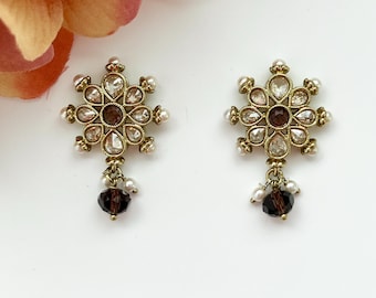 May include: A pair of gold-toned stud earrings. Each earring features a flower-shaped design with clear and brown crystals, small pearls, and a dangling dark brown bead. The earrings are set against a white background.
