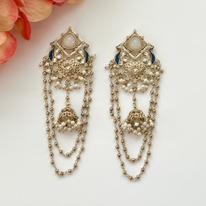 May include: A pair of ornate gold-toned earrings with a geometric design, featuring white stones, blue accents, and dangling pearl chains. The earrings have a bell-shaped charm at the bottom.