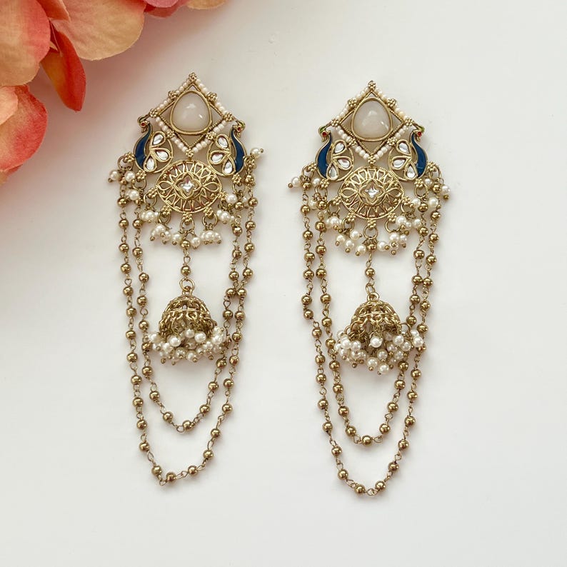 May include: A pair of ornate gold-toned earrings with a geometric design, featuring white stones, blue accents, and dangling pearl chains. The earrings have a bell-shaped charm at the bottom.
