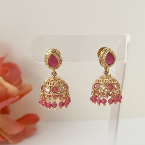 May include: A pair of gold-colored jhumka earrings. Each earring features a teardrop-shaped, ruby-colored stone at the top, with a filigree design. The bell-shaped bottom is adorned with small, ruby-colored beads and clear stones.