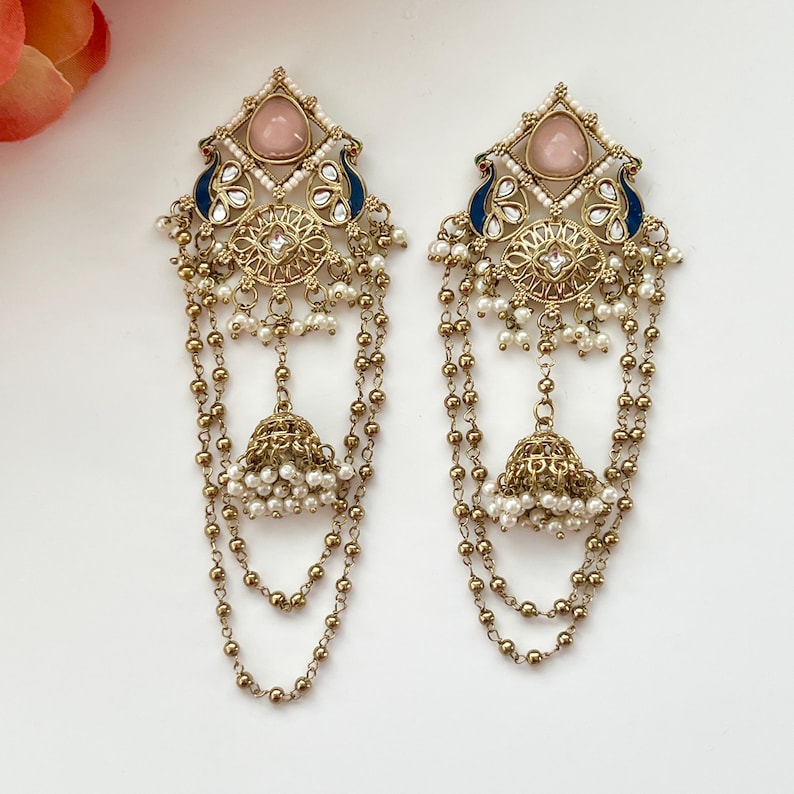 May include: A pair of ornate gold-toned earrings. Each earring features a pink gemstone, blue accents, and a peacock design. Dangling chains of beads and pearls lead to a bell-shaped charm. The earrings are designed for pierced ears.