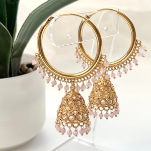 May include: Gold hoop earrings with dangling pink beads and ornate gold jhumka charms. The earrings are displayed on a clear acrylic stand.