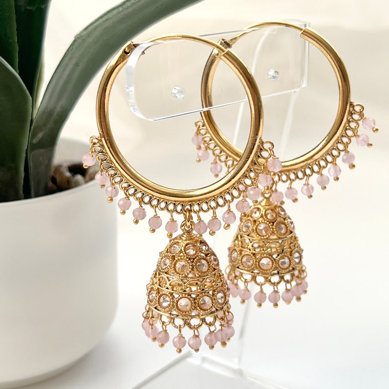 May include: Gold hoop earrings with dangling pink beads and ornate gold jhumka charms. The earrings are displayed on a clear acrylic stand.