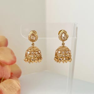 May include: A pair of gold-colored, ornate earrings. Each earring features a teardrop-shaped top and a bell-shaped bottom, adorned with small, clear stones. The earrings are displayed on a clear stand against a soft, neutral background.