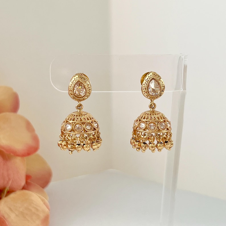 May include: A pair of gold-colored, ornate earrings. Each earring features a teardrop-shaped top and a bell-shaped bottom, adorned with small, clear stones. The earrings are displayed on a clear stand against a soft, neutral background.