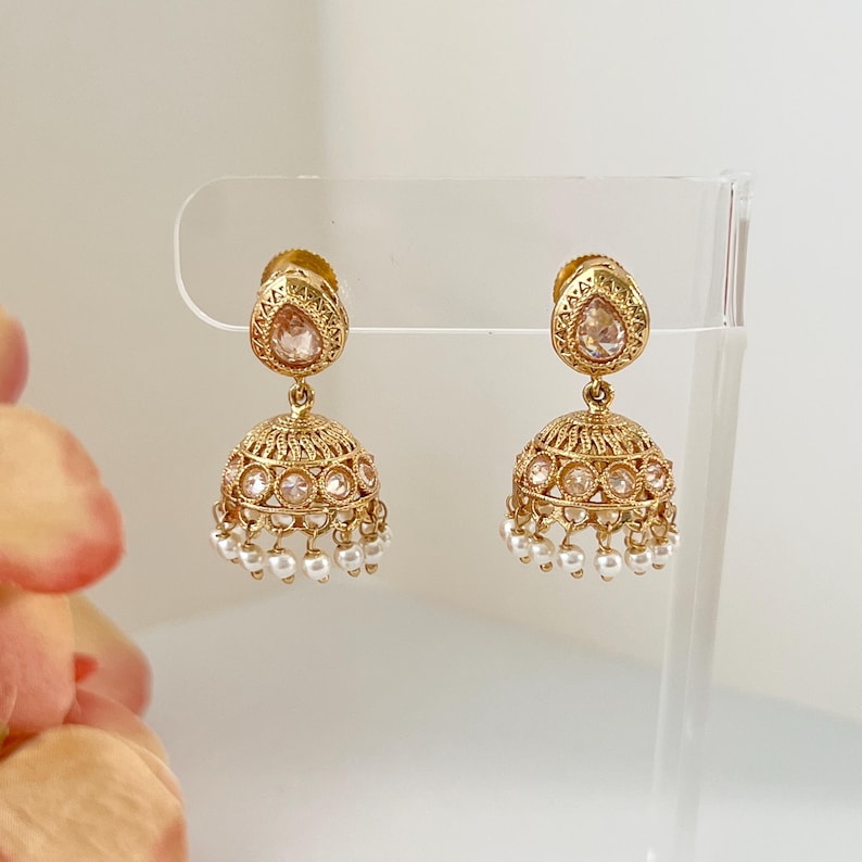 May include: A pair of gold-colored jhumka earrings. Each earring features a teardrop-shaped top with a pink gemstone and a bell-shaped bottom adorned with clear stones and small, dangling pearls. The earrings are displayed on a clear stand.