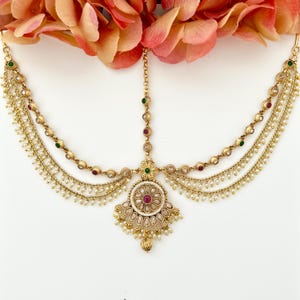 May include: A gold-toned headpiece with intricate detailing. The piece features a central pendant with a red gemstone surrounded by smaller white stones. Strands of pearls and small gold beads hang from the main structure, with red and green accents.