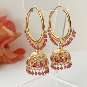 Könnte beinhalten: Goldfarbene Creolen mit einem baumelnden, glockenförmigen Anhänger. Die Ohrringe verfügen über rosa Edelsteine und kleine, baumelnde Perlen. Das Design ist aufwendig und detailliert, mit einem Fokus auf die filigrane Metallarbeit und den Kontrast von Gold und Rosa.