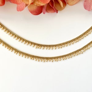 Pu&ograve; includere: Un paio di cavigliere dorate con delicati accenti di perle. Le cavigliere presentano un design a catena con piccole perle pendenti.