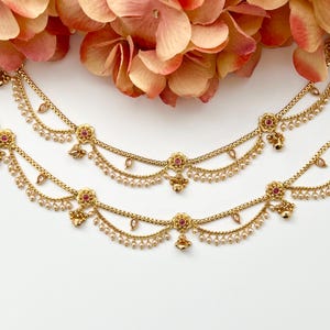 Pode incluir: Duas tornozeleiras douradas com detalhes de flores cor-de-rosa e gotas de p&eacute;rolas. As tornozeleiras est&atilde;o sobrepostas em um fundo branco.