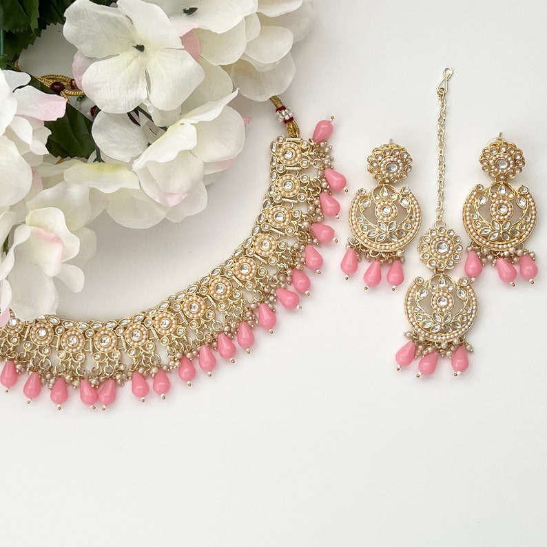 May include: A gold-toned jewelry set featuring a necklace, earrings, and a headpiece. The set is adorned with small white pearls and pink teardrop-shaped beads. White flowers are in the background.