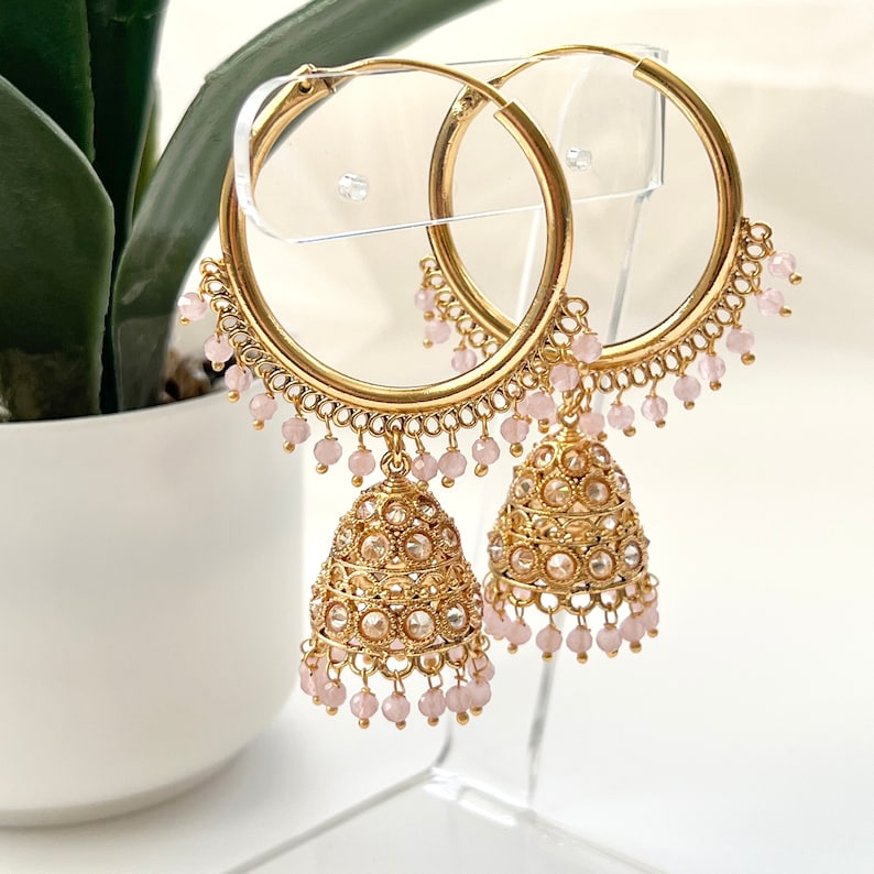 May include: Gold hoop earrings with dangling pink beads and ornate gold charms. The charms are decorated with clear stones.