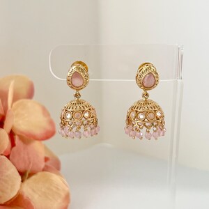 May include: A pair of gold-colored earrings with a teardrop-shaped pink stone at the top and a bell-shaped bottom. The earrings have a filigree design and small pink beads hanging from the bottom. The earrings are displayed on a clear stand.