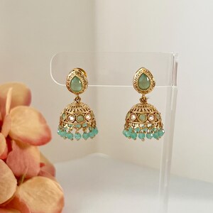 May include: A pair of gold-toned jhumka earrings. Each earring features a teardrop-shaped green stone at the top, a bell-shaped body with intricate detailing, and small turquoise beads dangling from the bottom. The earrings are displayed on a clear stand.