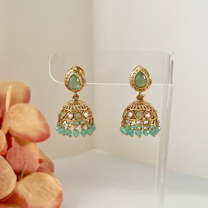 May include: A pair of gold-toned jhumka earrings. Each earring features a teardrop-shaped green stone at the top, a bell-shaped body with intricate detailing, and small turquoise beads dangling from the bottom. The earrings are displayed on a clear stand.