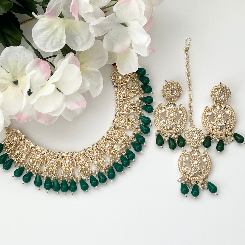 May include: A gold-toned necklace, earrings, and headpiece set with intricate detailing. The jewelry features small white pearls and teardrop-shaped emerald green stones. White flowers are in the background.