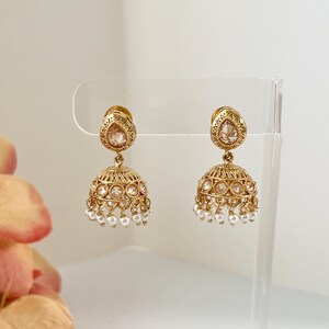 May include: A pair of gold-toned jhumka earrings. Each earring features a teardrop-shaped top with a central stone and a bell-shaped bottom adorned with small, round, white pearls. The earrings are displayed against a clear background.