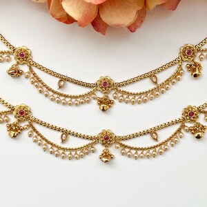 Pode incluir: Um par de tornozeleiras douradas com detalhes de flores cor-de-rosa e contas de p&eacute;rolas. As tornozeleiras t&ecirc;m um design de corrente delicado e sinos pendurados.
