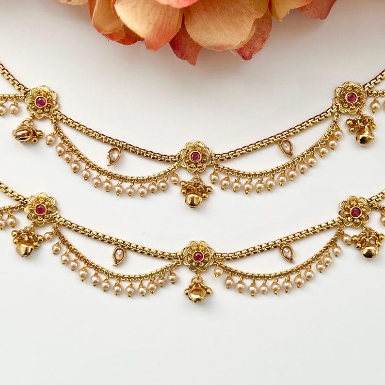 Pode incluir: Um par de tornozeleiras douradas com detalhes de flores cor-de-rosa e contas de p&eacute;rolas. As tornozeleiras t&ecirc;m um design de corrente delicado e sinos pendurados.