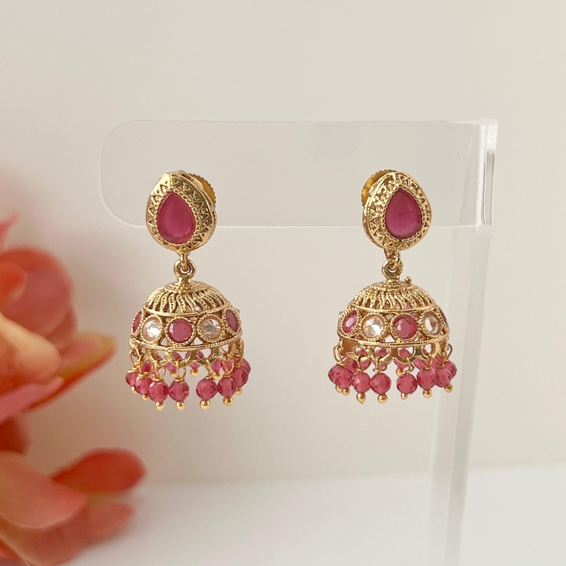 May include: A pair of gold-colored, ornate earrings. Each earring features a teardrop-shaped, magenta-colored stone at the top, with a bell-shaped design below. Small magenta beads dangle from the bottom of each earring.