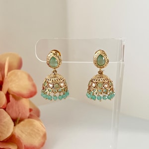 May include: A pair of gold-toned earrings with a bell-shaped design. Each earring features a teardrop-shaped, light green stone at the top and a cluster of small, light green beads at the bottom. The earrings are displayed on a clear stand.