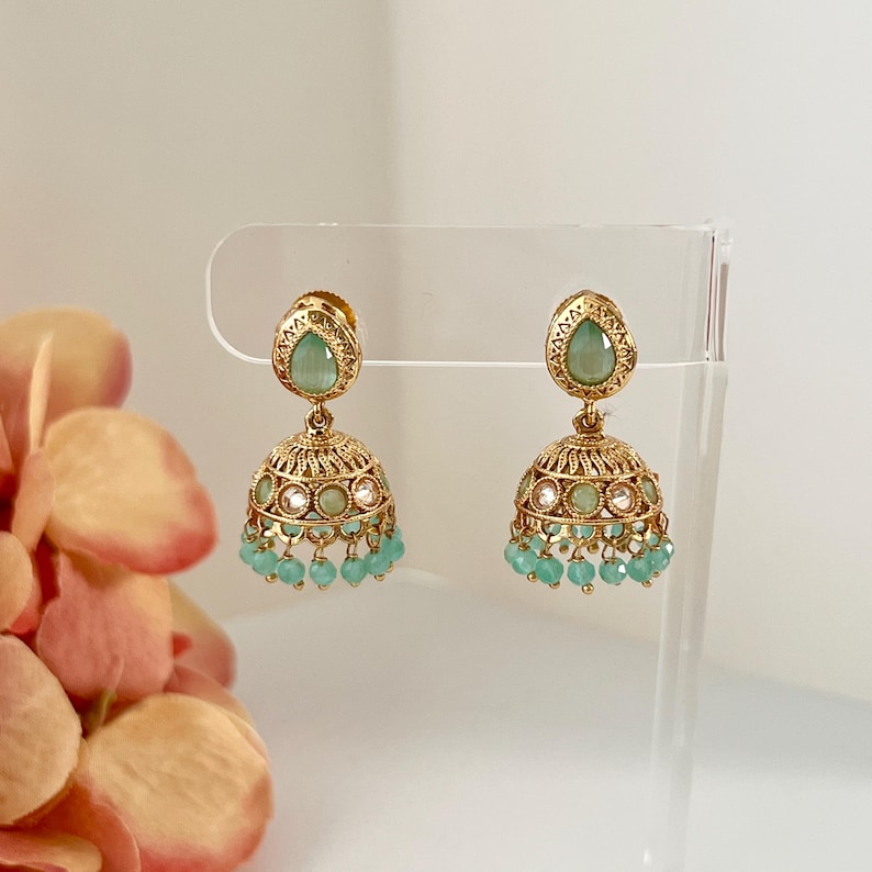 May include: A pair of gold-toned earrings with a bell-shaped design. Each earring features a teardrop-shaped, light green stone at the top and a cluster of small, light green beads at the bottom. The earrings are displayed on a clear stand.