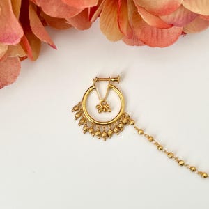 Op de afbeelding: Een goudkleurige neusring met een cirkelvormig ontwerp en bungelende kralenaccenten. De neusring heeft een kleine bloemenhanger en een delicate ketting van kleine kralen. De sieraden zijn geplaatst tegen een witte achtergrond met roze bloemenaccenten.