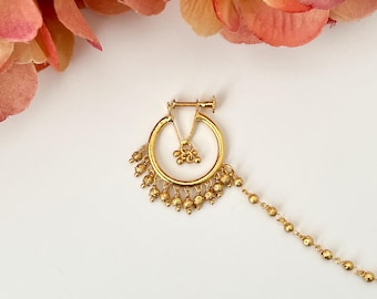 Puede incluir: Un aro de nariz dorado con un diseño circular y detalles colgantes de cuentas. El aro de la nariz presenta un pequeño colgante floral y una delicada cadena de pequeñas cuentas. La joya está colocada sobre un fondo blanco con detalles florales rosas.