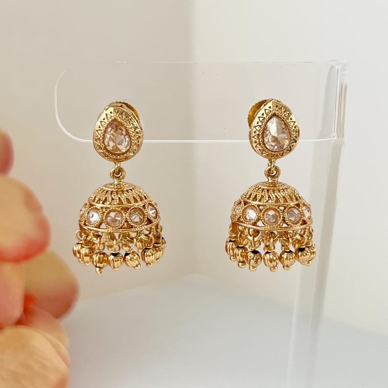 May include: A pair of gold-colored earrings with a teardrop-shaped top and a bell-shaped bottom. The earrings feature intricate detailing and small, clear stones. The bottom of the earrings have small, dangling gold beads.
