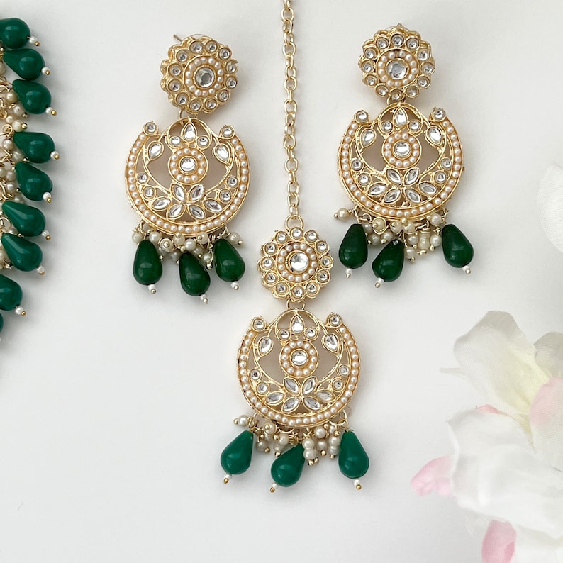 May include: Gold-toned jewelry set featuring a necklace, earrings, and a headpiece. The earrings and headpiece have a crescent design with white stones and small pearls, accented with green teardrop-shaped beads. The necklace has green beads.