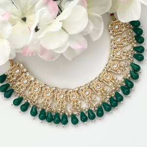 May include: A gold-toned necklace with a floral design, featuring small white pearls and clear crystals. The necklace has teardrop-shaped green beads hanging from the bottom. White and pink flowers are in the background.