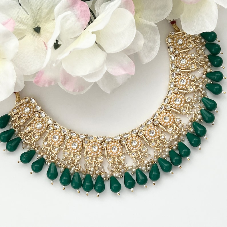 May include: A gold-toned necklace with a floral design, featuring small white pearls and clear crystals. The necklace has teardrop-shaped green beads hanging from the bottom. White and pink flowers are in the background.