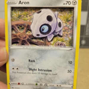 May include: A 3D shadow box trading card featuring the character Aron. The card displays 70 HP, the attacks Ram and Slight Intrusion, and text including No. 304 Iron Armor Pokemon, HT 1'4, WT 132.3 lbs, and the copyright 2023 Pokemon Creatures GAME FREAK.