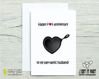 May include: A white greeting card featuring a black heart-shaped skillet illustration in the center. The text reads: Happy iron heart anniversary to my pan-tastic husband! The card is paired with a matching white envelope, presenting a clean design.